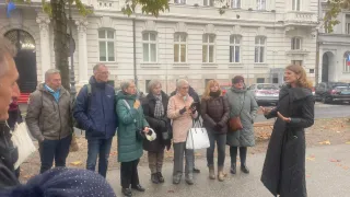 Eine Gruppe von Menschen steht vor einem großen weißen Gebäude. Sie sind alle warm gekleidet. Eine Frau spricht zu der Gruppe.