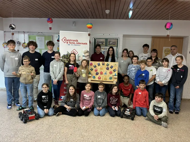 Eine Gruppe von Schülern steht um ein Display-Board mit verschiedenen Pins und Abzeichen herum. Sie stehen und sitzen auf dem Boden und tragen Freizeitkleidung. Einige Schüler halten kleine Roboter.