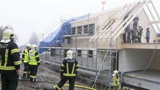 A firefighter with a helmet and reflective vest stands on a building under construction, surrounded by scaffolding. Other firefighters are nearby, with one holding a hose.