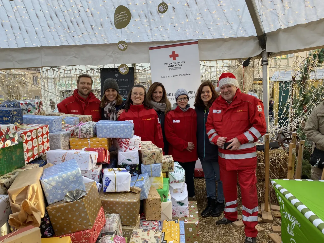 A group of smiling volunteers, dressed in red, stand behind a table full of wrapped gifts. They are under a tent with a banner that reads Osterreichisches Rotes Kreuz.