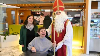 Ein Mann, der als St. Nikolaus verkleidet ist, steht neben einem lächelnden Rollstuhlfahrer, mit einer Frau im Hintergrund.