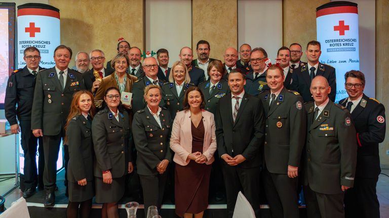 Eine Gruppe von Militärangehörigen und Zivilbeschäftigten steht lächelnd vor einem dekorierten Hintergrund. Einige tragen formelle Kleidung, während andere Uniformen tragen. Eine Frau in einer rosa Jacke steht in der Mitte.