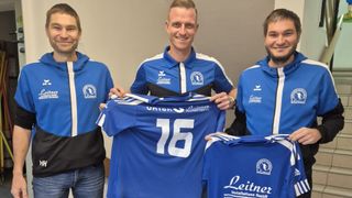 Drei Männer halten ein blaues Fußballtrikot mit der Nummer 76 und lächeln für ein Foto. Der Mann in der Mitte trägt ein blaues Shirt mit Logo. Die Männer tragen blaue Hoodies und Hosen.