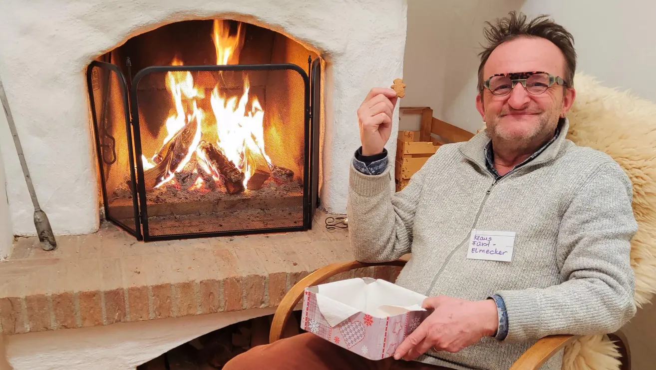 A man with glasses and a sweater holds a gingerbread and a box of tissues in front of a fireplace.