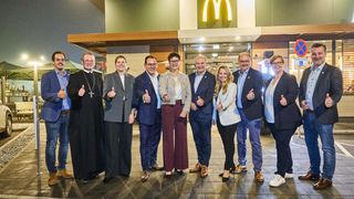 Eine Gruppe formell gekleideter Personen steht vor einem McDonald's und gibt Daumen hoch. Hinter ihnen ist das beleuchtete McDonald's-Schild zu sehen.