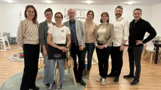 A group of people, including a woman in a white shirt and a man in glasses, stand in a room. They are posing for a picture in front of a wall with the text 'LITTI's Kindercafe.' There is a rug on the floor.