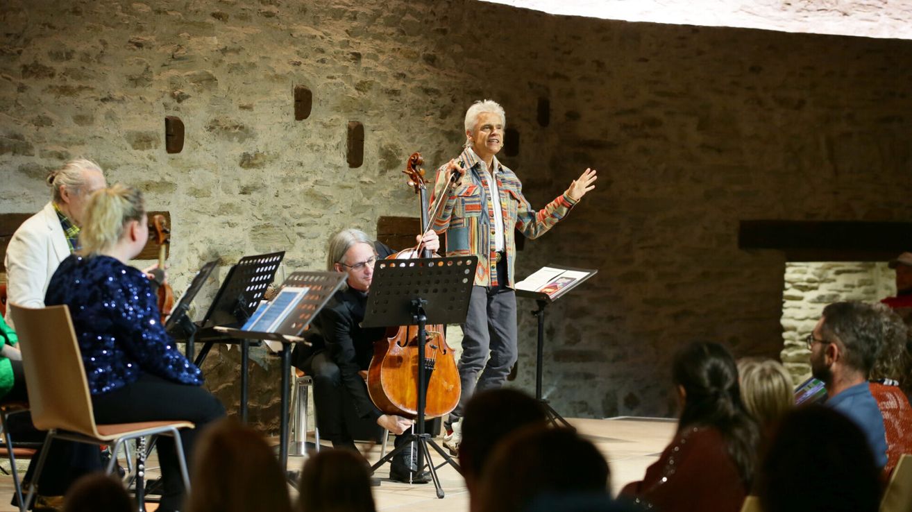 Ein Musiker spielt auf der Bühne mit einem Cello. Ein anderer Musiker sitzt hinter ihm, und Notenständer sind aufgestellt. Das Publikum sitzt vor ihnen.