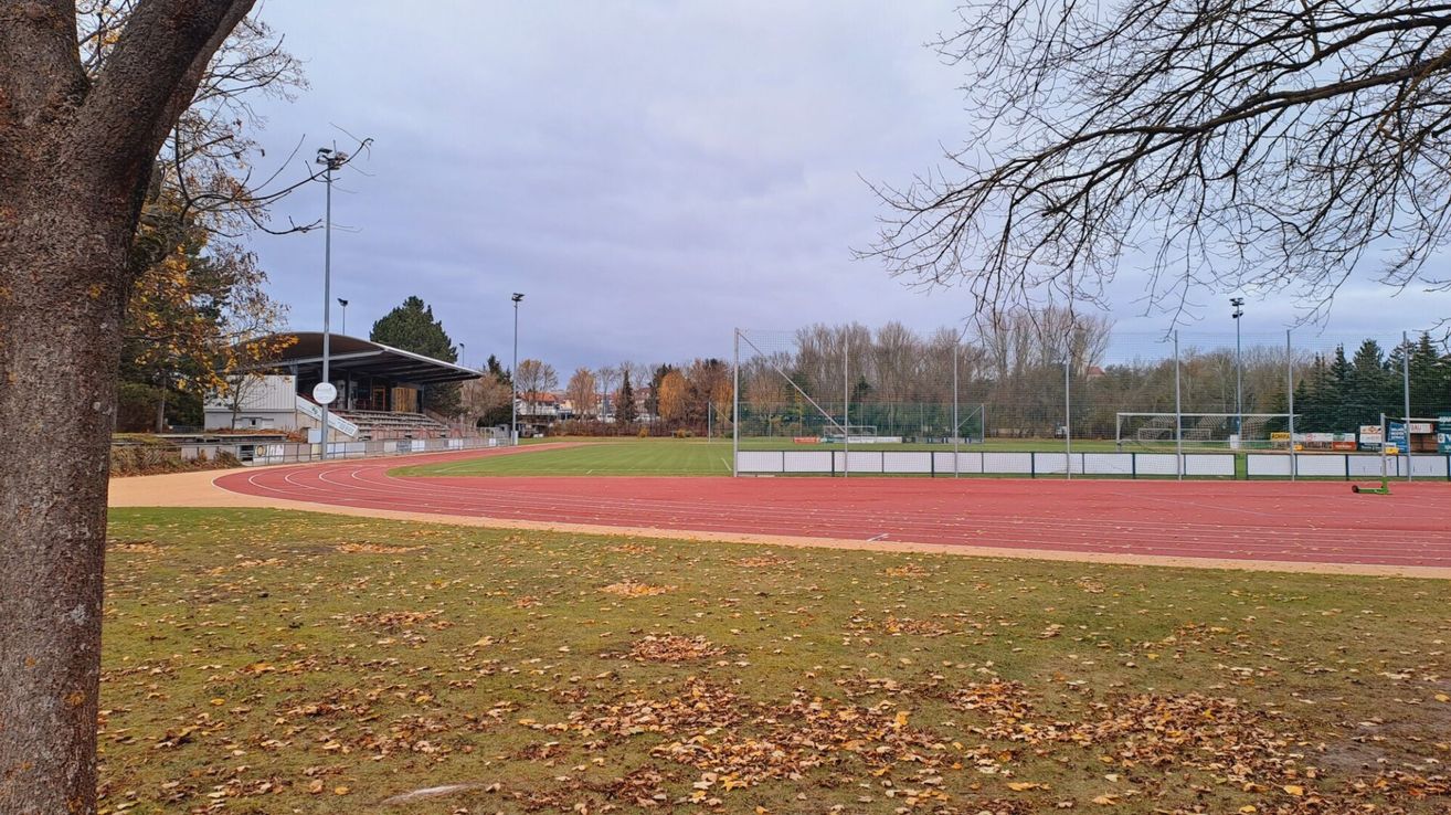 Ein Freiluft-Sportstadion mit einer leeren roten Laufbahn, umgeben von einem Grasfeld und Bäumen. Straßenlaternen sind auf der linken Seite sichtbar.