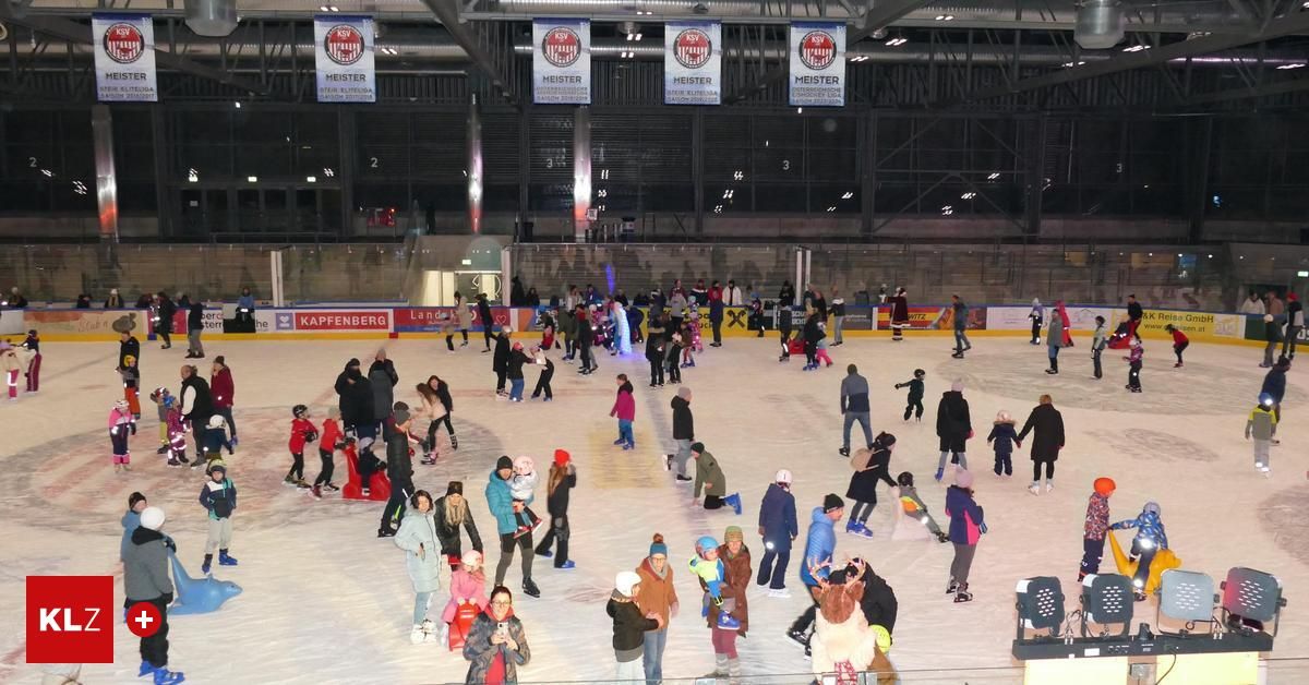 An indoor ice skating rink with multiple banners. Many people in winter clothing skate on the ice, while others stand around watching. Some are holding children, and others are in groups. The rink has a modern ceiling with lights.