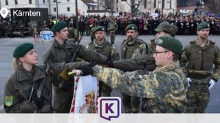Soldaten in Tarnuniformen halten eine Flagge während einer Zeremonie vor einer Menge, mit Gebäuden und Bäumen im Hintergrund.