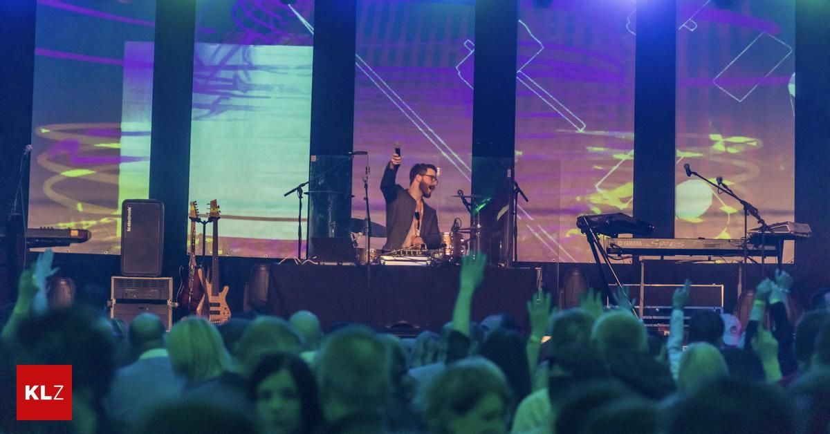 Ein Mann steht mit einem Mikrofon auf der Bühne, vor einem Publikum, das die Hände hebt. Hinter ihm stehen eine Gitarre, Trommeln und ein Keyboard. Ein großer Bildschirm mit bunten Grafiken ist im Hintergrund.