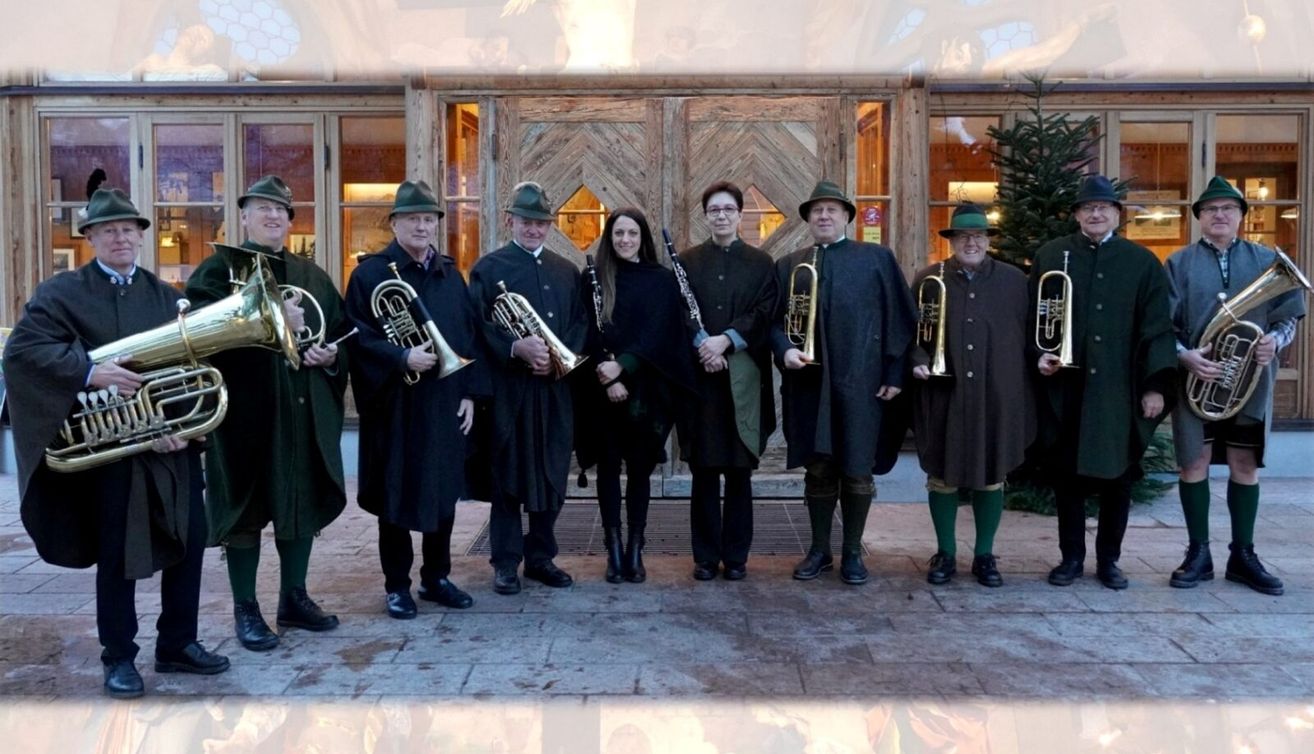 Eine Gruppe von Musikern steht vor einem Holzgebäude. Sie halten Instrumente in den Händen und lächeln. Die Frau in der Mitte trägt ein schwarzes Oberteil und eine Hose.