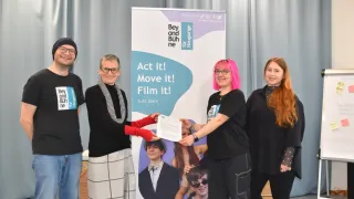 Vier Personen stehen vor einem Banner mit Text und Bildern. Sie halten ein Papier und lächeln. Das Banner sagt 'Act it! Move it! Film it!' und zeigt ein Foto von zwei Kindern. Sie tragen Freizeitkleidung.