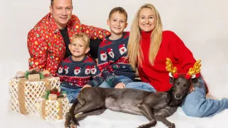 Eine Familie zu viert sitzt mit ihrem Hund zusammen, alle in Weihnachtspullovern gekleidet. Der Mann, die Frau und die beiden Jungen lächeln, während der Hund Hirschgeweih trägt. Geschenke sind vor ihnen platziert.