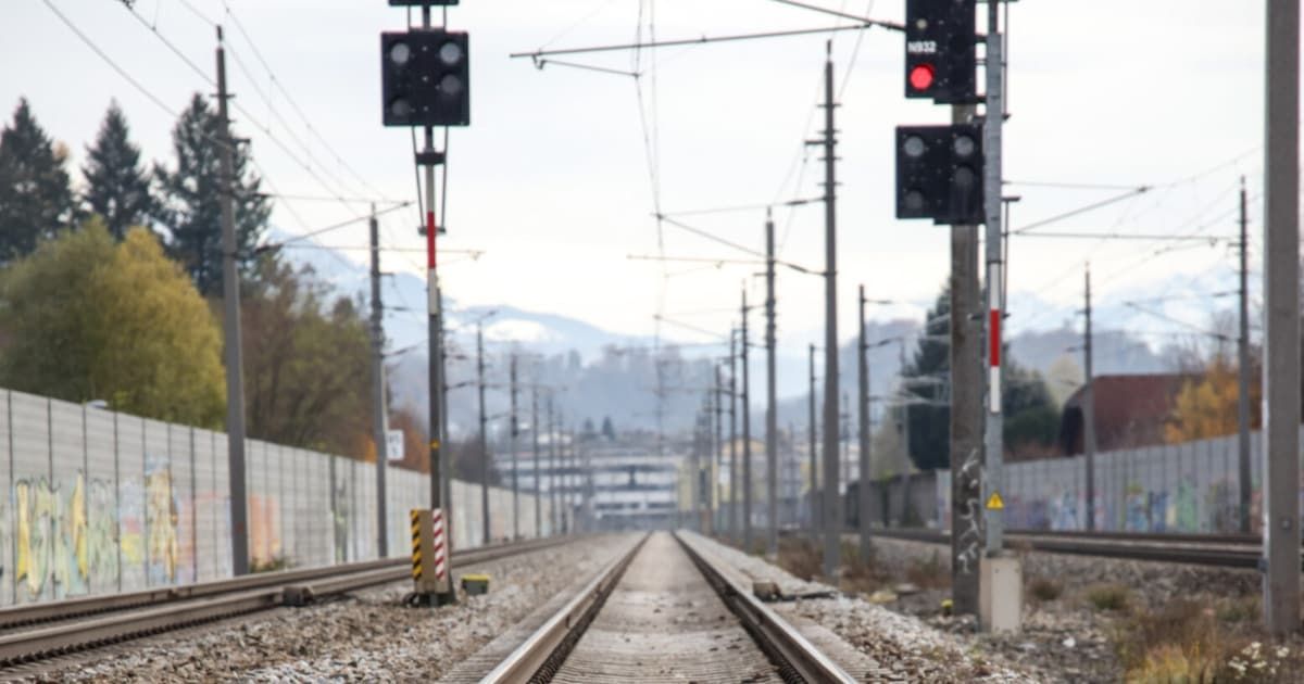 A railroad track with a red signal light. The track is empty, and there are utility poles and wires on either side. In the distance, there are buildings, mountains, and trees.