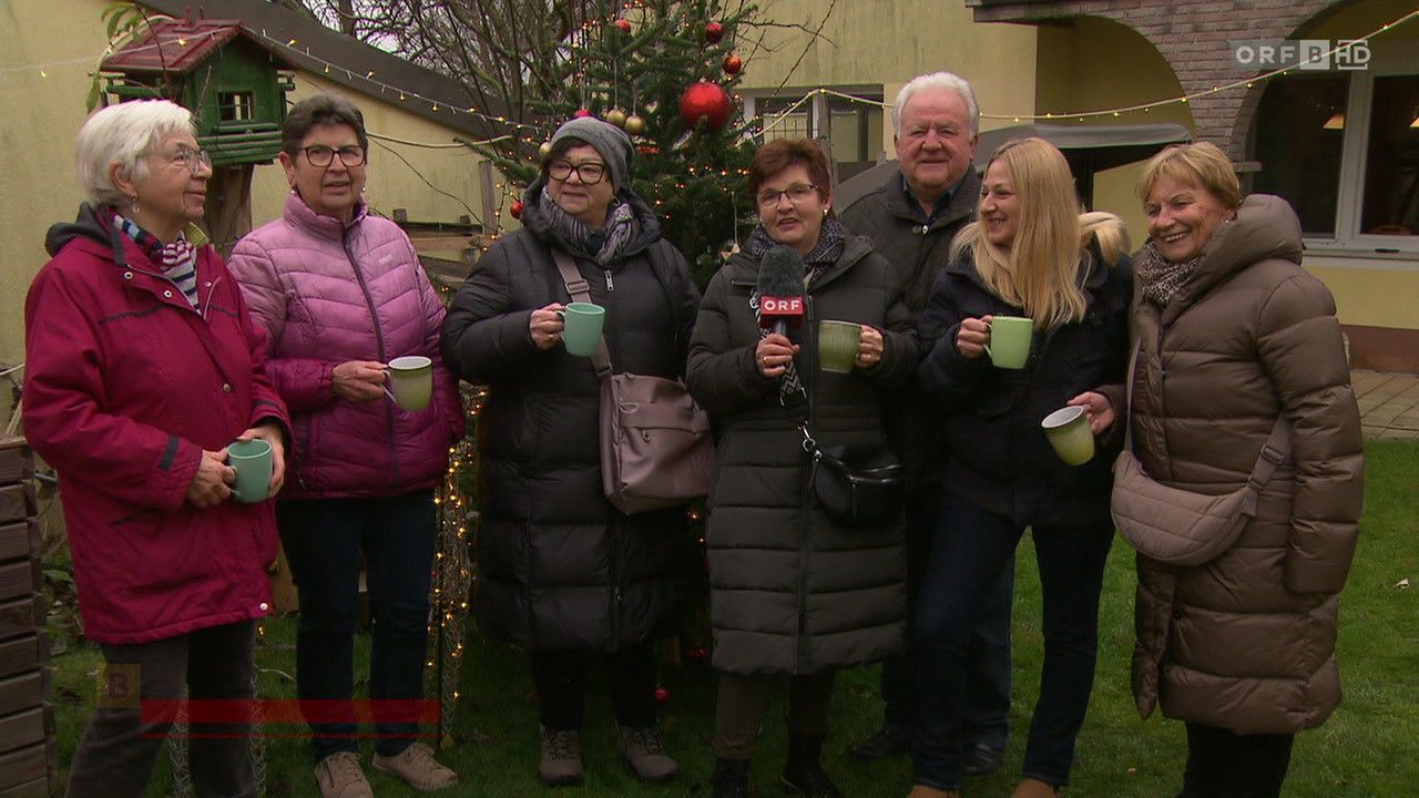 Fünf Erwachsene stehen vor einem geschmückten Weihnachtsbaum, lächeln und halten grüne Tassen. Eine Frau in der Mitte hält ein Mikrofon. Sie sind in Winterkleidung gekleidet.