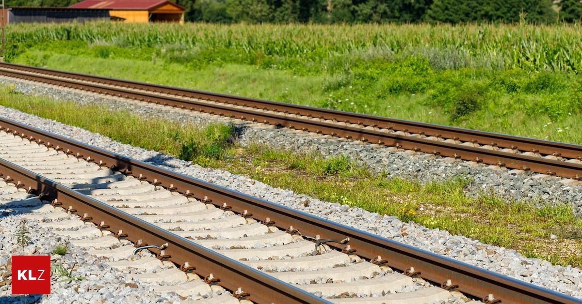 Two railway tracks with gravel on the sides. A small house is visible in the distance. The background features a vast green field.
