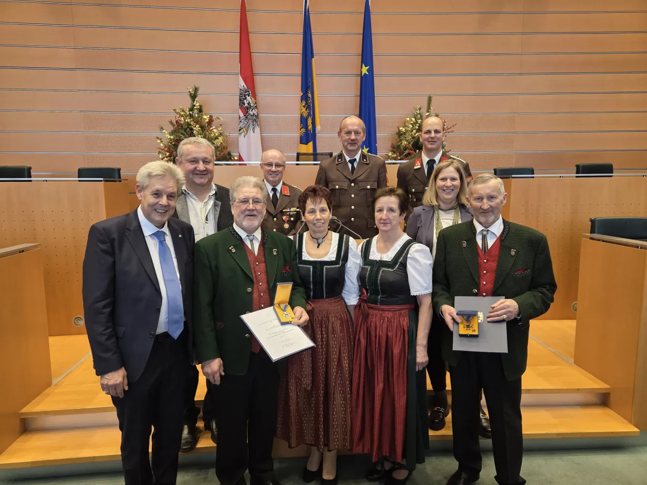 Group photo of people in formal attire with medals, standing on a stage with flags behind them.