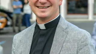 A smiling priest in glasses and a gray suit poses outside a building with parked cars and people in the background.