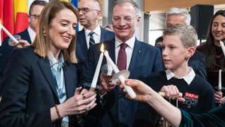 Eine Frau und ein Mann in formeller Kleidung halten brennende Kerzen, die von einem jungen Jungen angezündet werden. Dahinter stehen andere Menschen in formeller Kleidung, darunter Brillen und Krawatten.