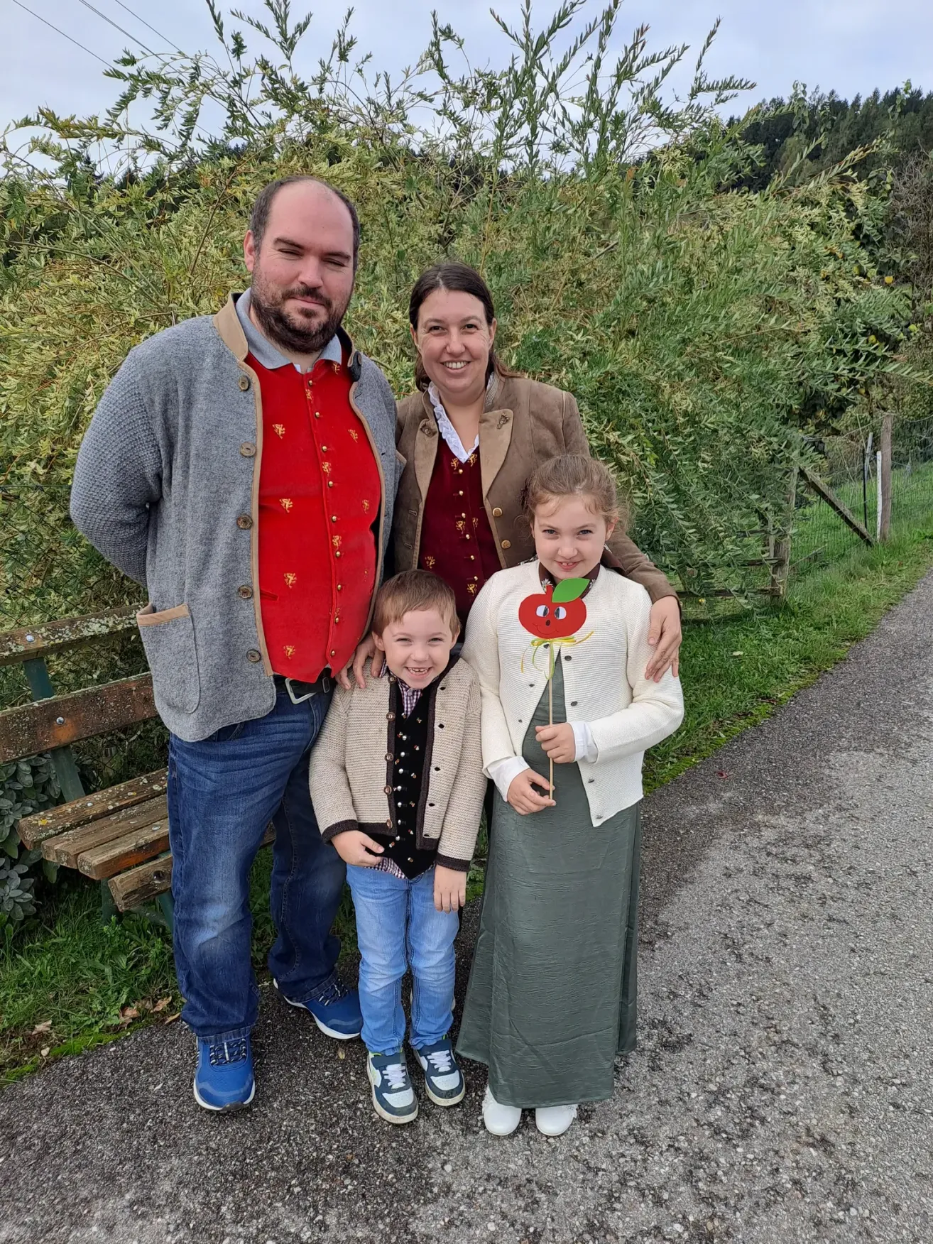 A family of four poses for a photo on a sunny day in a park. The father, mother, and two children stand close together. The children are wearing jackets, and the mother has an apple decoration on her sweater.