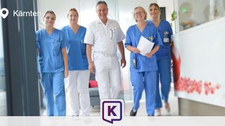 Fünf medizinische Fachkräfte in blauen Kitteln gehen durch einen Krankenhauskorridor. Eine Frau hält eine Mappe.