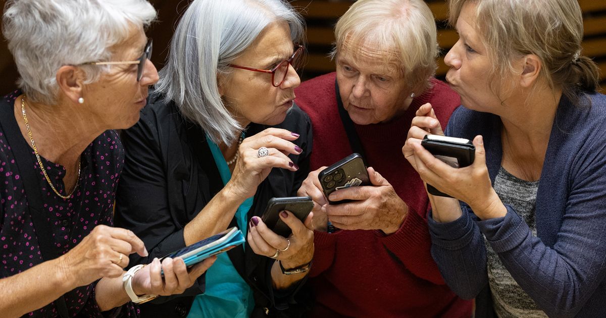 Four women, likely seniors, are engrossed in their smartphones, possibly discussing a message or app. They are seated together, with one holding a wallet.