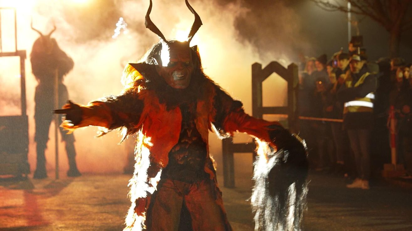 Eine Person in einem Hörnerkostüm mit einem orangefarbenen und schwarzen Outfit steht auf einer Bühne vor einer Holzstruktur. Die Person hält ein schwarzes Tuch in der linken Hand. Das Gesicht der Person wird von Licht beleuchtet.