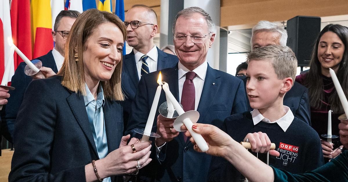 Eine Frau und ein Mann, beide mit brennenden Kerzen, lächeln einen jungen Jungen an. Dahinter schauen ein weiterer Mann und eine Frau, einer mit Brille, zu. Flaggen im Hintergrund.