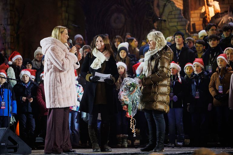 Drei Frauen stehen auf der Bühne, eine hält ein Mikrofon und spricht, während eine andere Blumen und ein Papier hält. Hinter ihnen steht eine Menschenmenge, einige mit Mützen.