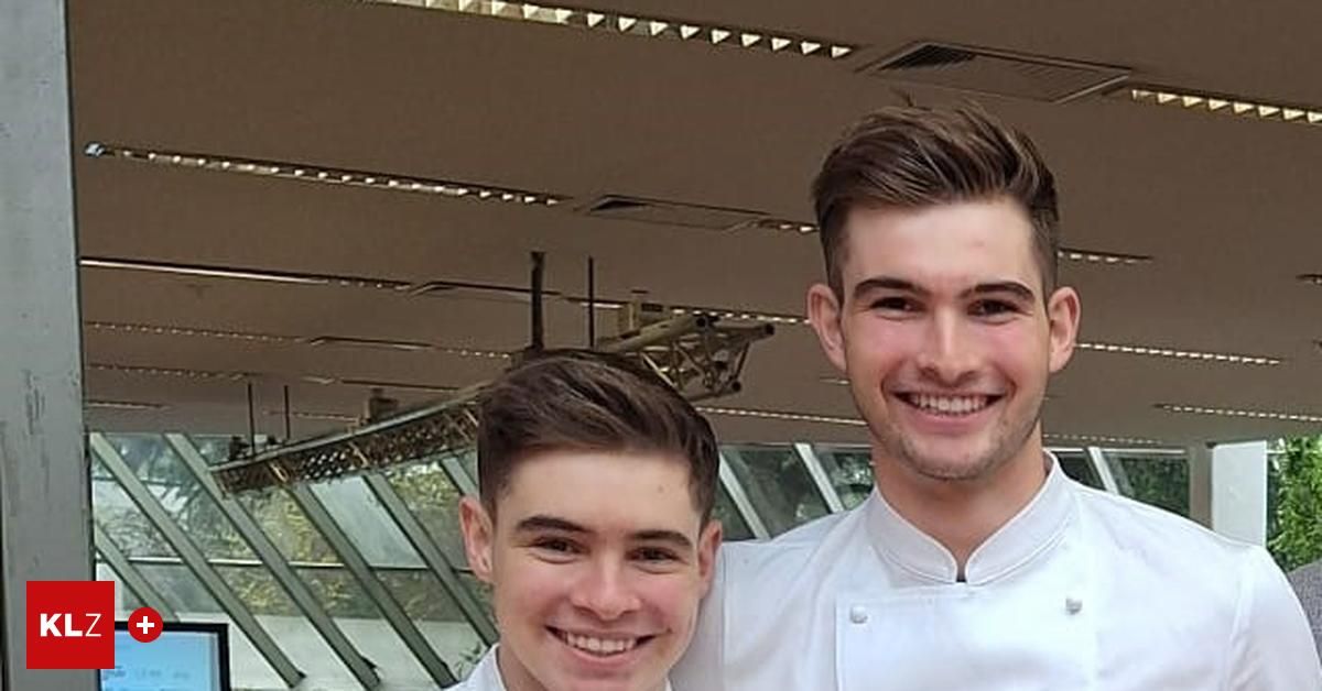 Zwei junge Männer in Kochuniformen stehen lächelnd nebeneinander. Der Mann auf der linken Seite hat kurze Haare und trägt ein weißes Hemd. Der Mann auf der rechten Seite hat einen Bart und trägt eine Kochjacke. Hinter ihnen ist ein großes Fenster.