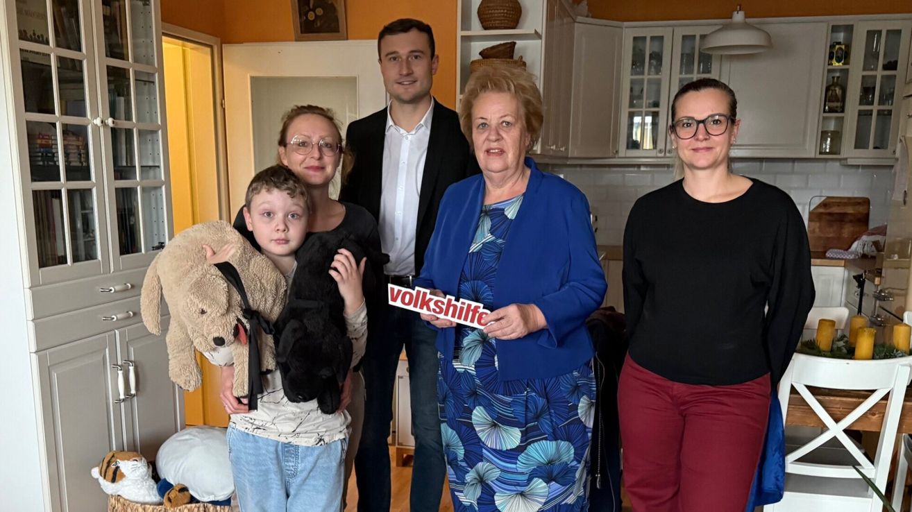 Eine Familie mit einem Kind und einer Frau, die ein Schild mit der Aufschrift 'volkshilfe' hält. Sie sind in einem Raum mit einem lächelnden Gesicht.