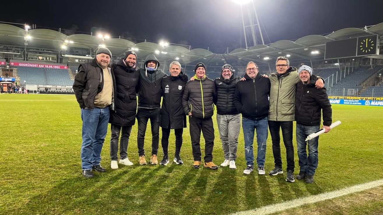 Eine Gruppe von Männern steht auf einem Fußballfeld und trägt Winterkleidung. Sie lächeln und posieren für ein Foto.