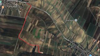 Luftaufnahme eines ländlichen Gebiets mit verschiedenen Feldern, einer Straße namens Hauptstraße und einer roten Linie, die einen Pfad oder eine Grenze anzeigt. Die Landschaft umfasst grüne und braune Flecken.