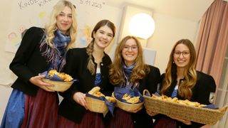 Vier Frauen, jede mit einer blauen Schleife, stehen zusammen und halten Körbe mit Gebäck. Sie lächeln und posieren für ein Foto vor einer weißen Wand mit Datum und Licht.