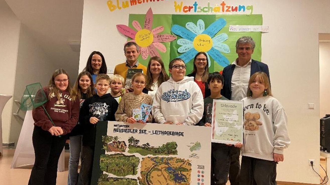 Eine Gruppe von Menschen, einschließlich Kindern, steht vor einem farbenfrohen Hintergrund mit Blumen und Text. Sie halten eine Karte und Urkunden in den Händen. Einige von ihnen tragen eine Brille.