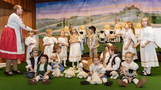 Kinder in traditionellen Kostümen führen ein Krippenspiel mit Engeln, Hirten und Schafen vor einem gemalten Hintergrund auf.