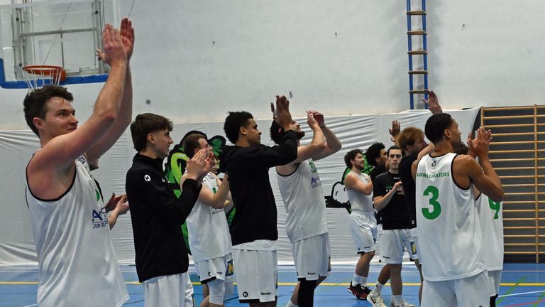 Eine Gruppe Basketballspieler in weißen und schwarzen Trikots feiert auf einem Spielfeld. Hände sind zum Sieg erhoben, und ein Banner ist im Hintergrund sichtbar.