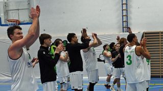 Eine Gruppe Basketballspieler in weißen und schwarzen Trikots feiert auf einem Spielfeld. Hände sind zum Sieg erhoben, und ein Banner ist im Hintergrund sichtbar.
