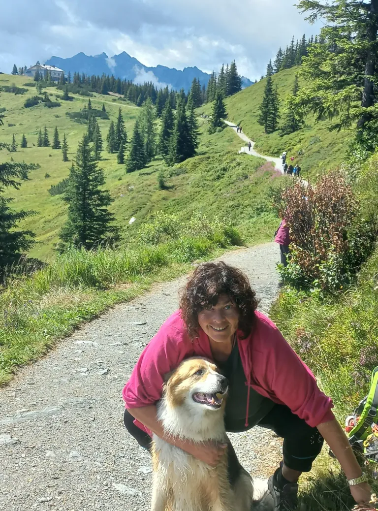 Eine Frau mit lockigen Haaren lächelt und hockt sich hin, um mit ihrem Hund auf einem Bergpfad zu posieren.
