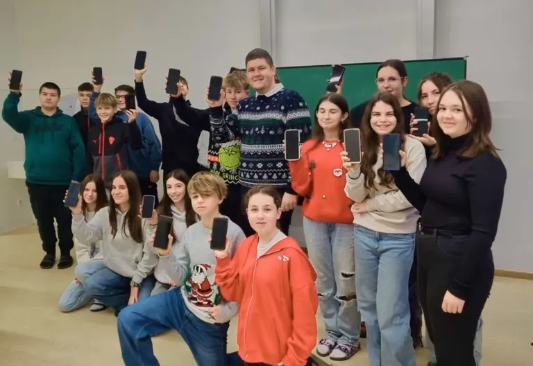 Eine Gruppe von Teenagern posiert für ein Foto, alle halten Smartphones, einige tragen festliche Kleidung. Ein Teenager in der Mitte trägt einen Pullover.