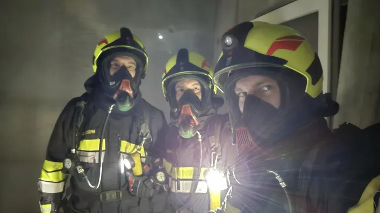 Drei Feuerwehrleute in voller Ausrüstung, mit gelben Helmen mit roten und schwarzen Akzenten, posieren für ein Foto mit einem hellen Licht, das den Vordergrund beleuchtet.