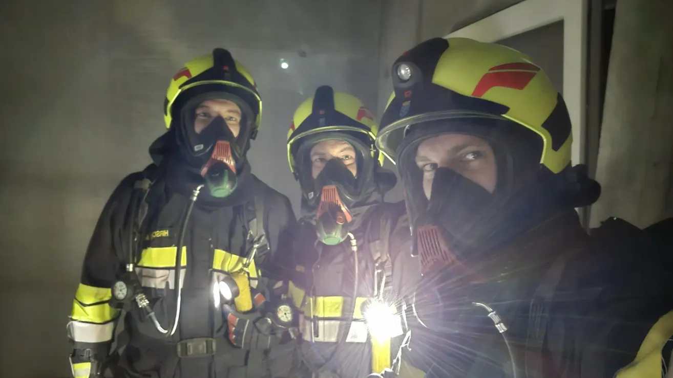 Drei Feuerwehrleute in voller Ausrüstung, mit gelben Helmen mit roten und schwarzen Akzenten, posieren für ein Foto mit einem hellen Licht, das den Vordergrund beleuchtet.