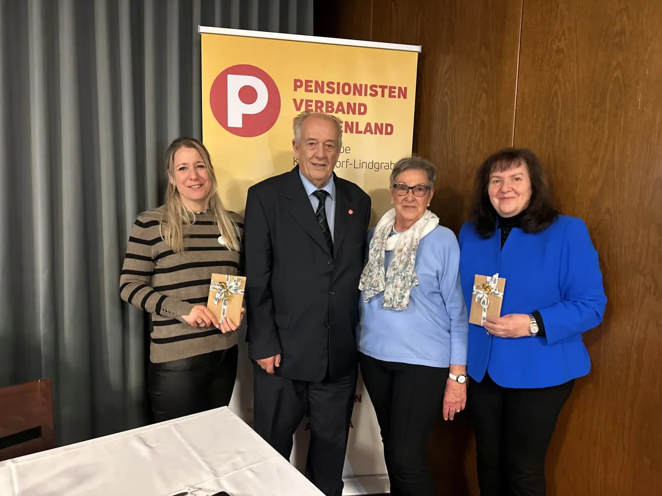 Eine Gruppe von vier Erwachsenen steht vor einem Banner mit dem Text 'Pensionisten Verband'. Der Mann trägt einen Anzug, und die Frauen tragen Brillen und Schals. Sie alle lächeln und halten ein Geschenk. Hinter ihnen steht ein Tisch mit weißer Tischdecke.
