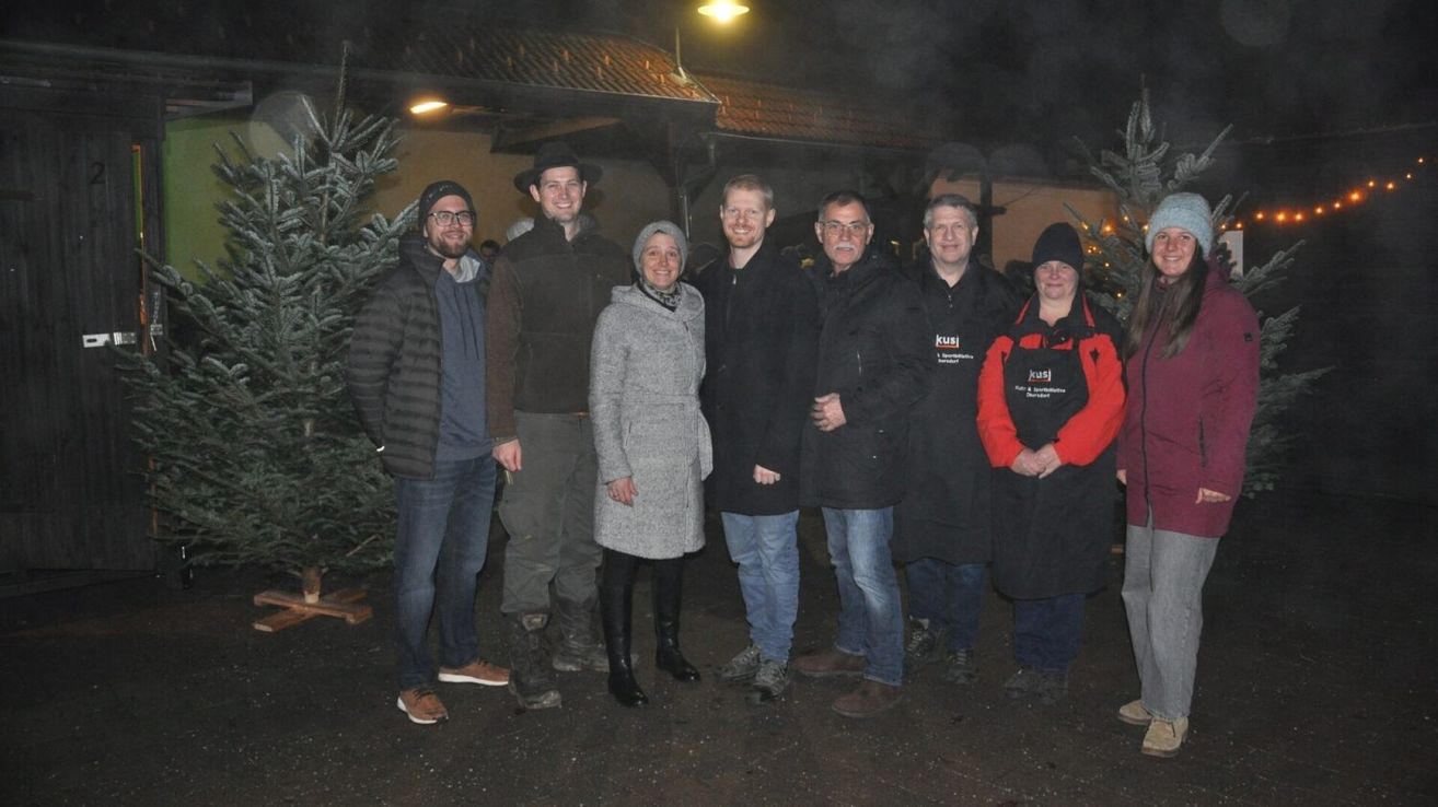 Eine Gruppe von Menschen steht draußen vor Tannenbäumen. Sie sind in Winterkleidung gekleidet, einige tragen Mützen. Die Frau in der Mitte trägt eine Schürze. Der Mann auf der rechten Seite hat ein Logo auf seiner Jacke. Der Hintergrund ist schwach beleuchtet, was auf Nachtzeit hindeutet.