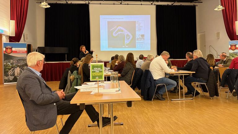 Eine Frau spricht in ein Mikrofon vor einer Projektionsleinwand. Zuhörer sitzen um Tische herum und lauschen. Ein grünes Schild sagt 'Für ein klimafreundliches Leben.'