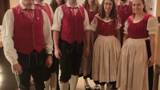 Eine Gruppe von Menschen in traditioneller Kleidung, die lächelnd für ein Foto in einem Raum posiert. Sie stehen eng beieinander, mit einer Holzwand und einer herzförmigen Dekoration im Hintergrund.