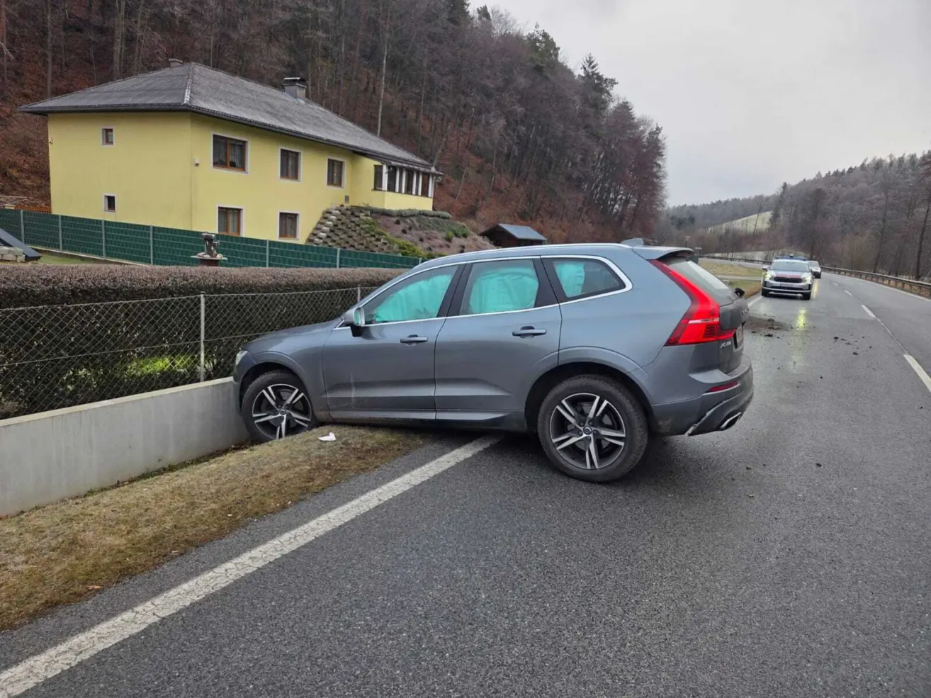Ein grauer SUV ist am Straßenrand geparkt, mit der Heckpartie auf einem Grasstreifen. Ein gelbes Haus befindet sich hinter einem Zaun in der Nähe. Ein anderes Auto fährt in die entgegengesetzte Richtung.