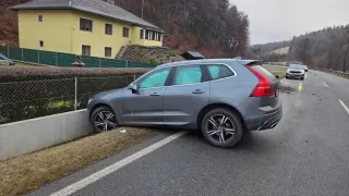 Ein grauer SUV ist am Straßenrand geparkt, mit der Heckpartie auf einem Grasstreifen. Ein gelbes Haus befindet sich hinter einem Zaun in der Nähe. Ein anderes Auto fährt in die entgegengesetzte Richtung.