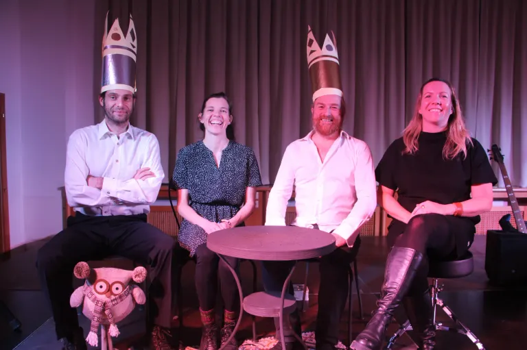 Vier Personen sitzen um einen kleinen Tisch herum und tragen Papierkronen. Sie scheinen für ein Foto zu posieren und lächeln.